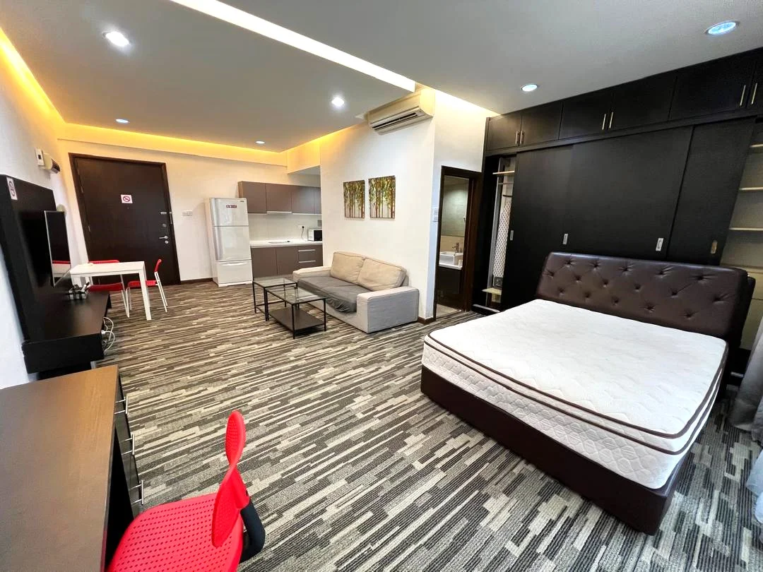 Room features: queen-size bed, upholstered headboard, sofa in grey fabric, coffee table, TV console, wall-mounted TV, desk with chair, white painted walls, recessed downlights, laminate flooring, built-in wardrobe, kitchenette with white cabinetry, stainless-steel fridge, microwave, sink, bathroom visible in background; style: modern.